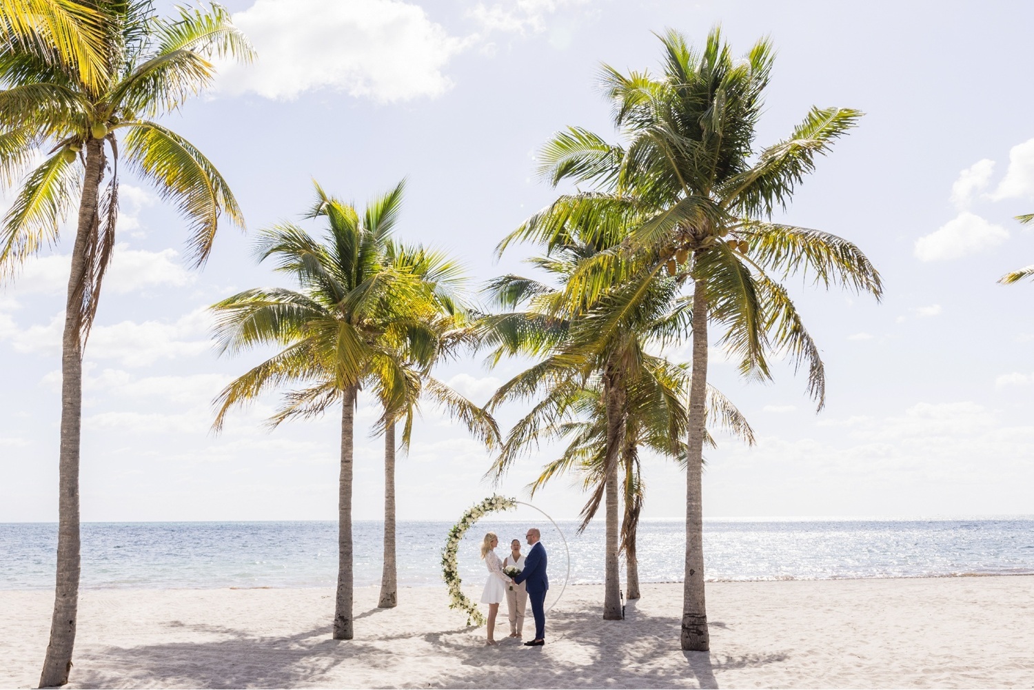 Beach wedding ceremony setup with white chairs facing a floral-draped arch between palm trees, overlooking the ocean.