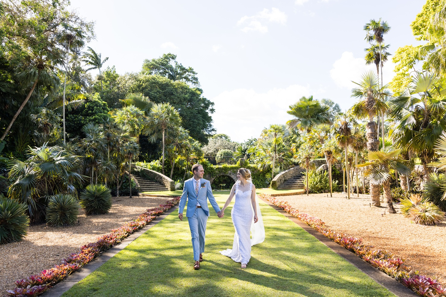 Outdoor wedding ceremony in a tropical garden, with the couple standing on a grassy aisle surrounded by palm trees and seated guests.