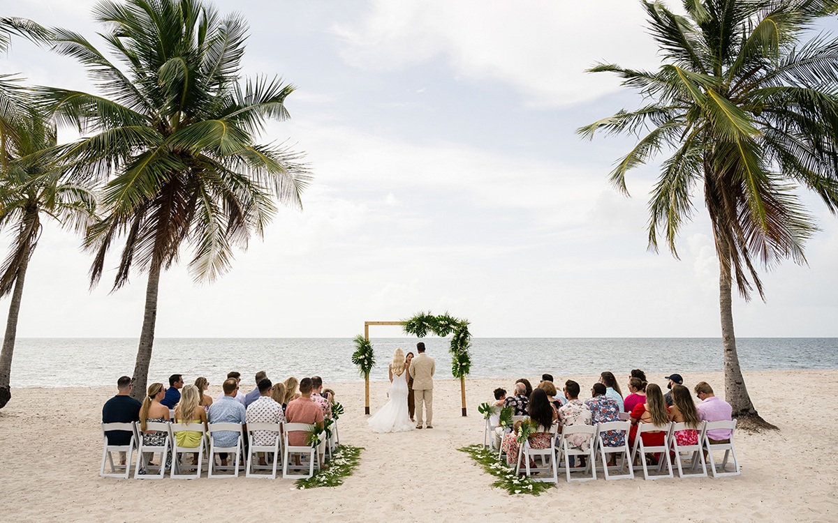 Bride and groom kissing under a wooden wedding arch decorated with white fabric and floral arrangements on a waterfront deck.